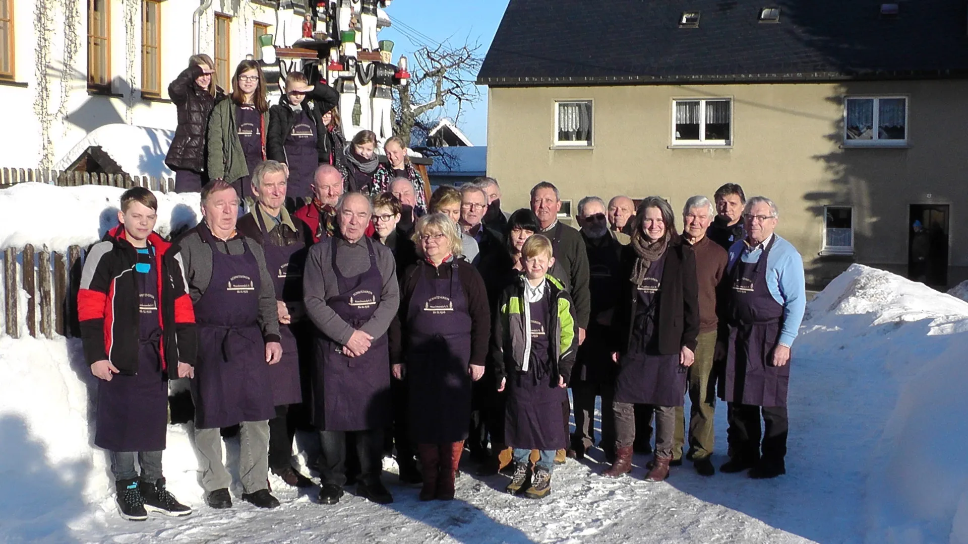 Gruppenfoto der Vereinsmitglieder in Schnitzerschürzen im Schnee vor der Ortspyramide