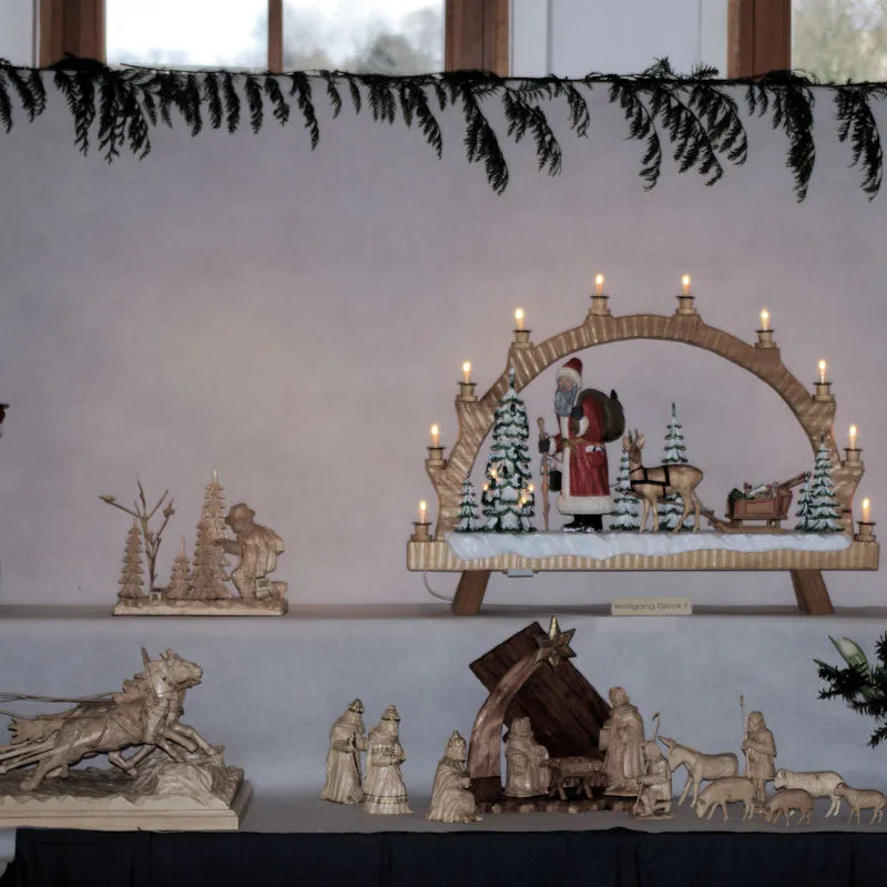 Schwibbogen mit geschnitztem Weihnachtsmann, Rentier und Schlitten im verschneiten Wald, davor Krippenfiguren und ein Pferdegespann